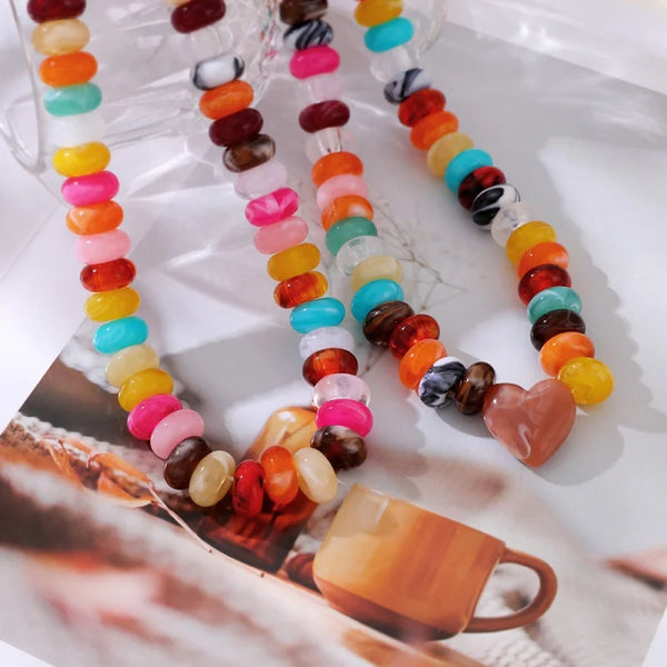 A close-up of several colorful beaded necklaces arranged on a glossy white surface with a printed coffee cup graphic. The necklaces feature round and marbled beads in bright shades of orange, yellow, pink, turquoise, brown, black, and white, with one necklace including a heart-shaped bead.