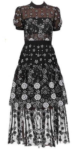Zeze is a sheer mesh black midi cocktail dress. Short puff sleeve with white flower embroidery detail. A-line with stand, round neck layers of lace and large frills. 